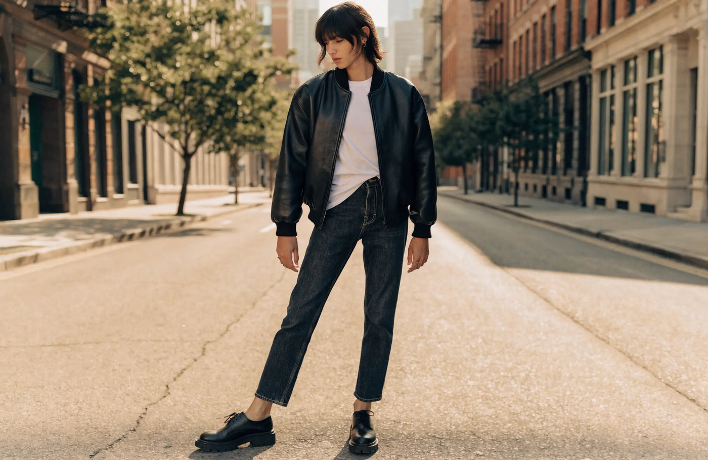Person wearing a black leather bomber jacket with a white t-shirt and jeans on a city street