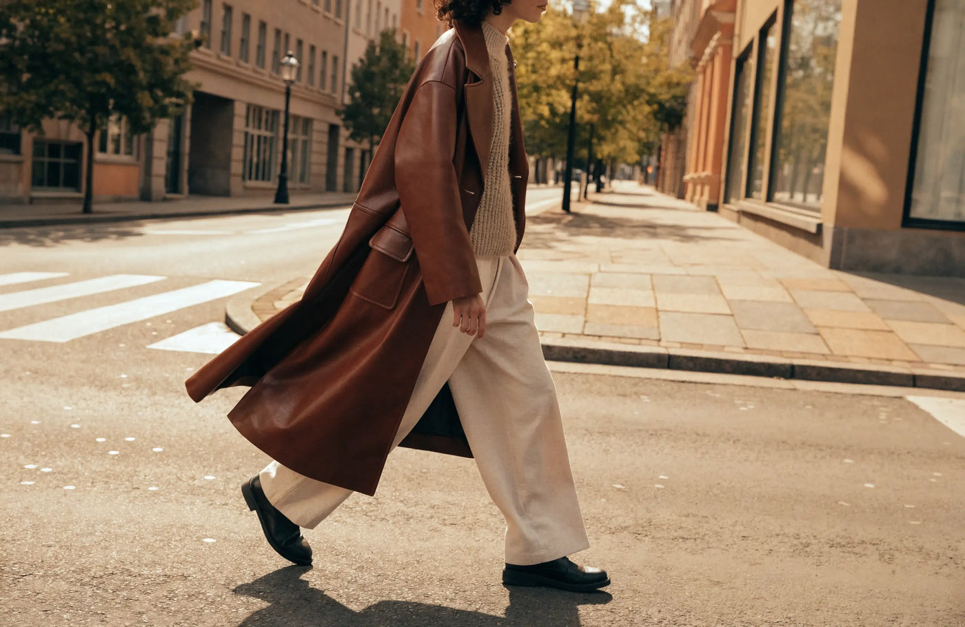 Person wearing a long brown leather coat over a knit sweater on an autumn city street